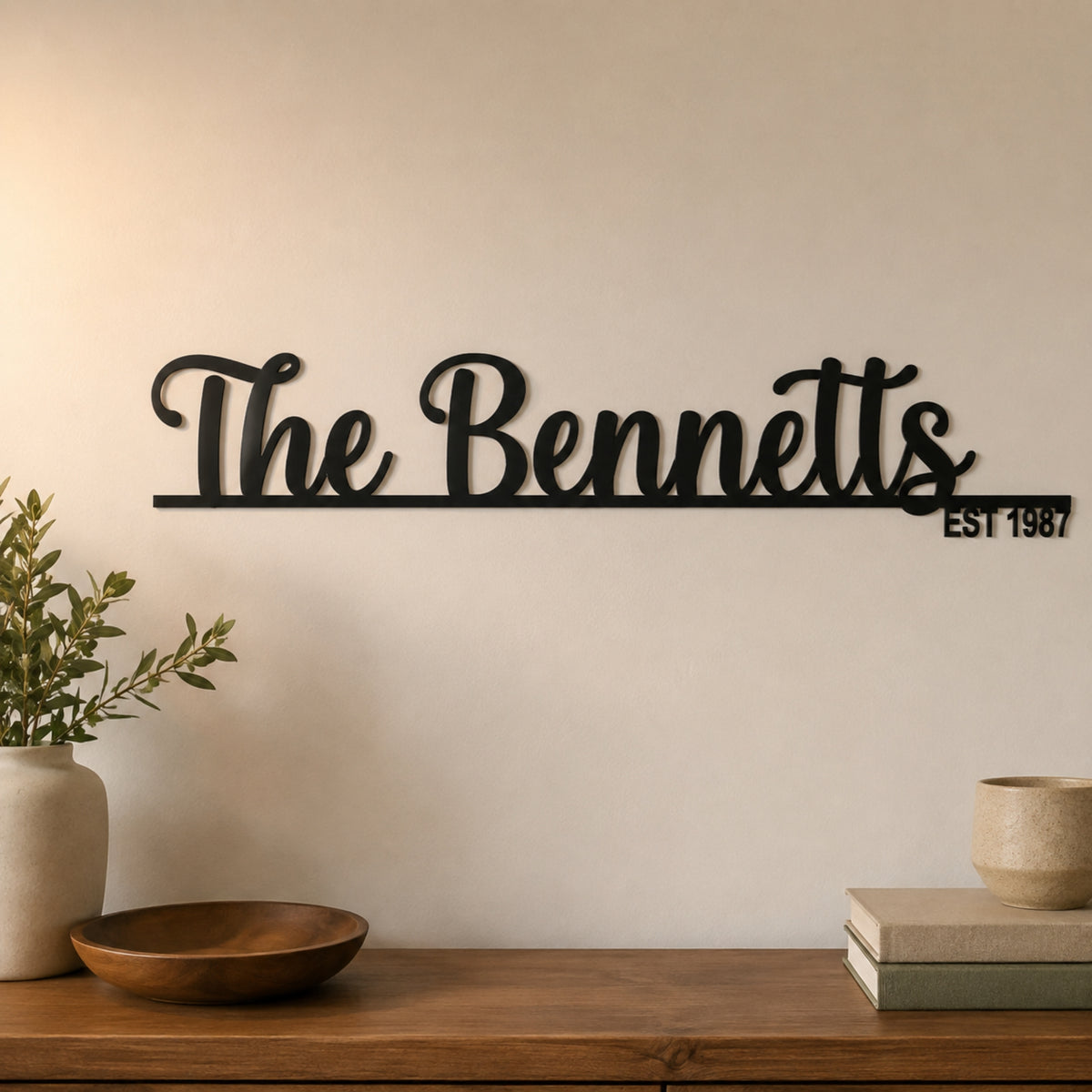 Black metal script wall sign reading 'The Bennetts' with 'EST 1987' on a horizontal bar, mounted on a beige wall above a wooden console table with decorative objects. Main image
