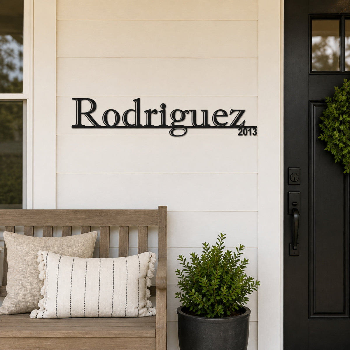 Custom metal house sign displaying 'Rodriguez 2013' in black script and numerals, mounted on a white shiplap wall on a front porch next to a wooden bench and potted plant. Main image