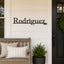 Custom metal house sign displaying 'Rodriguez 2013' in black script and numerals, mounted on a white shiplap wall on a front porch next to a wooden bench and potted plant.