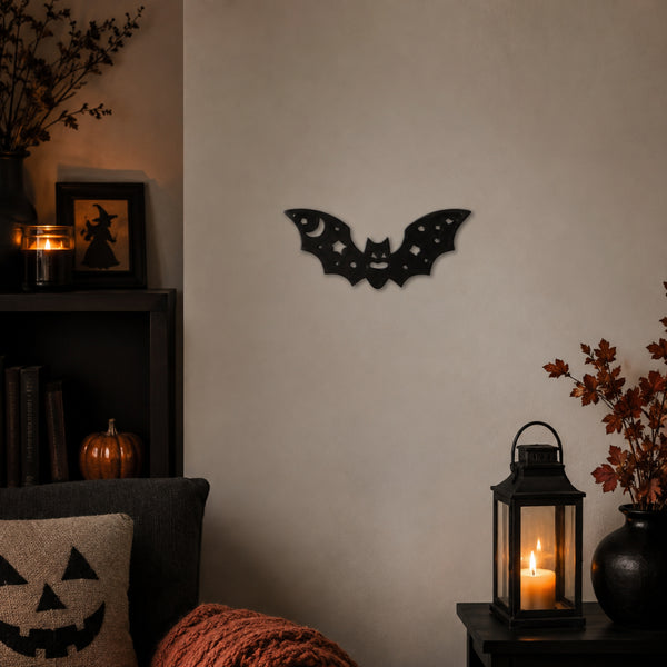 A black bat-shaped wall decor piece with a crescent moon and stars cutout, mounted on a beige wall in a cozy, dimly lit room with candles and autumn decor.