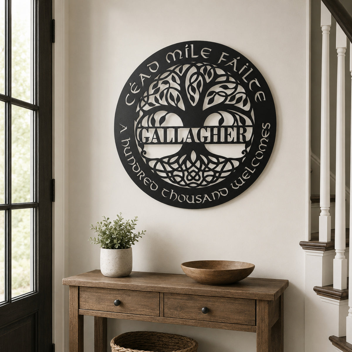 A circular black metal wall sign featuring a tree of life design and the text 'CæD mIle FÁlCe' and 'A honored thousand welcomes' around the edge, mounted on a beige wall above a wooden console table in a home interior.