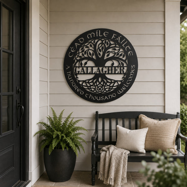 A circular black metal wall sign with a tree of life design and the text 'CÉAD MILE FÁILTE GALLACHER' and 'HUNDRED THOUSAND WELCOMES' is mounted on a beige siding wall on a porch. A black bench with cream and beige pillows and a throw blanket sits below, next to a potted fern.