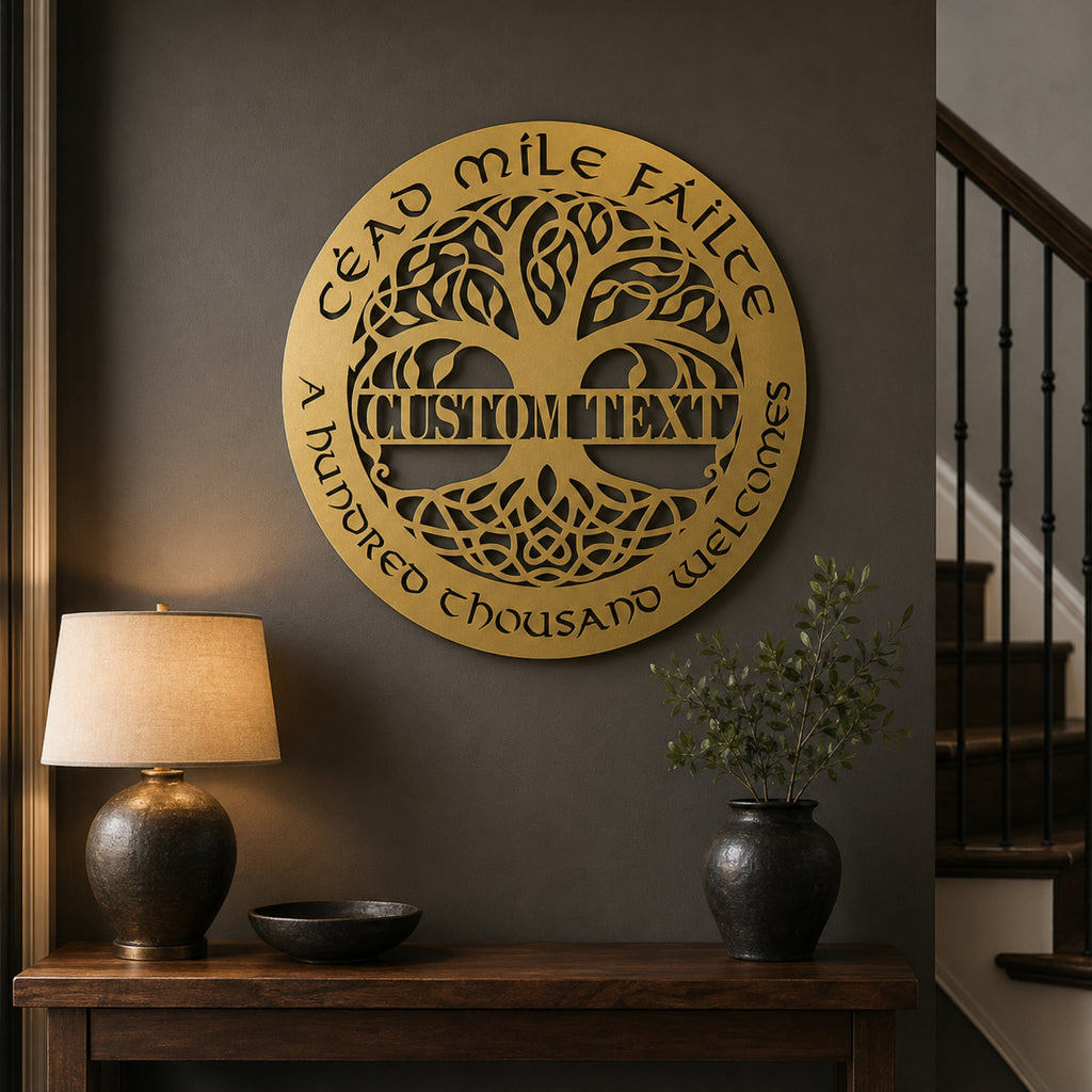 A round gold metal wall sign with a tree of life design and customizable text, mounted on a dark gray wall above a wooden console table with a lamp and plant.