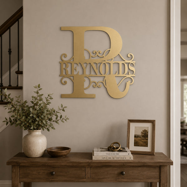 A large decorative gold metal wall sign spelling 'Reynolds' in an ornate monogram style, mounted on a beige wall above a wooden console table with a vase, bowl, books, and a framed picture.