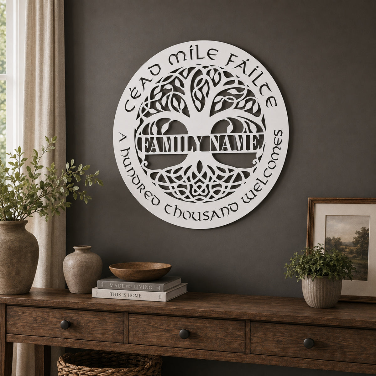 A round white metal tree-of-life wall sign with a personalized name in the center and the words 'A hundred thousand welcomes' and 'Dear mile family' around the edge, displayed on a dark gray wall above a wooden console table with vases, books, and a framed picture.