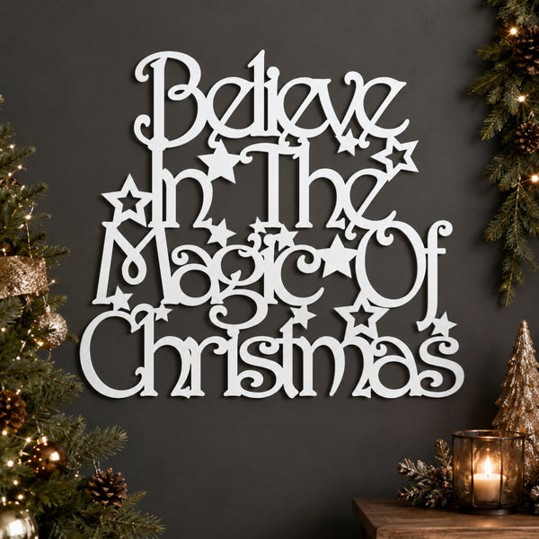 White laser-cut wooden wall art with the phrase 'Believe in the Magic of Christmas' surrounded by stars, displayed on a dark gray wall with Christmas tree branches and a candle in the background.