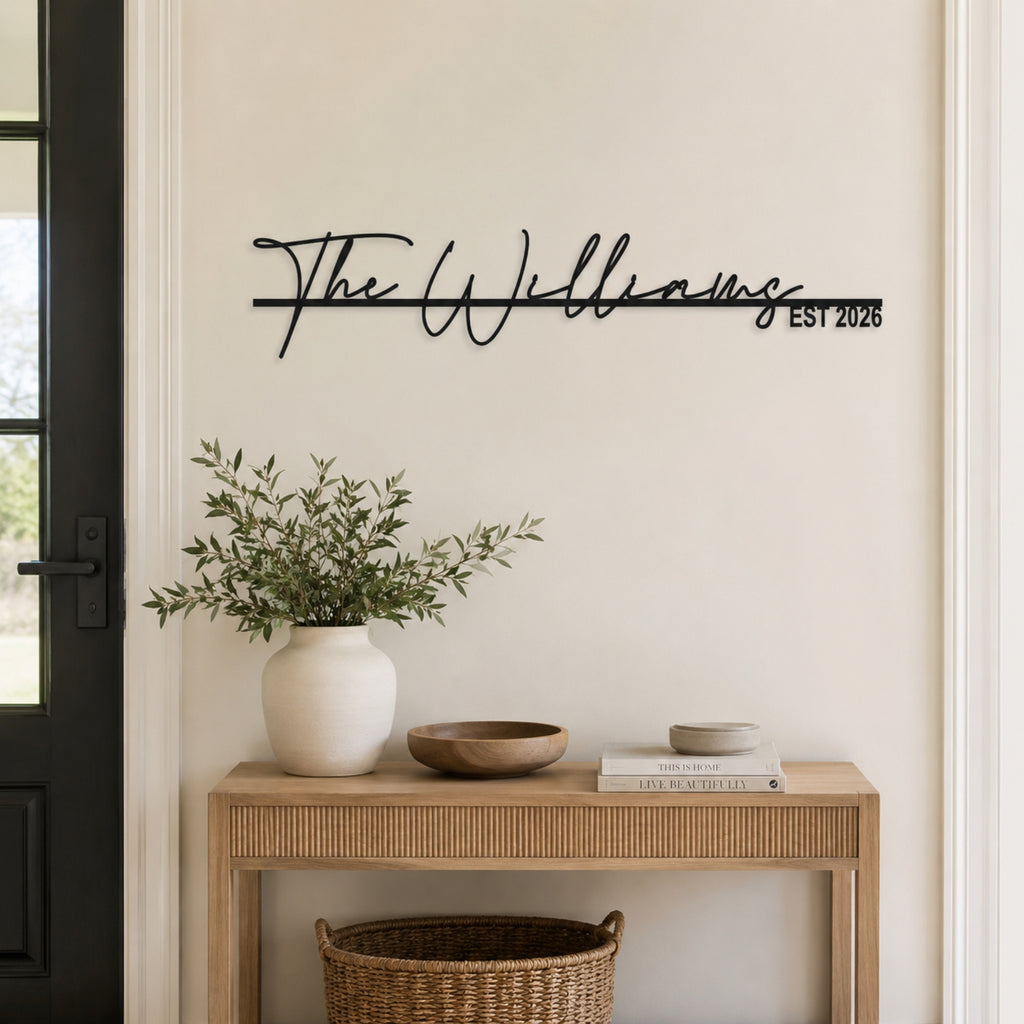 Black metal personalized wall art featuring the name 'The Williams' in script with 'EST 2026' on a horizontal line, mounted on a beige wall above a wooden console table with a white vase of greenery.