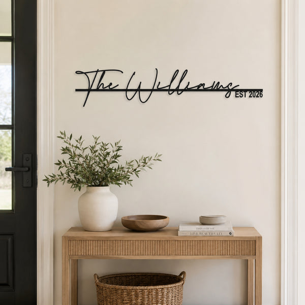 Black metal personalized wall art featuring the name 'The Williams' in script with 'EST 2026' on a horizontal line, mounted on a beige wall above a wooden console table with a white vase of greenery.