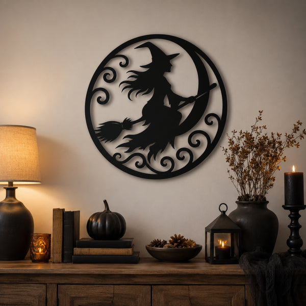 Black metal wall art featuring a witch riding a crescent moon with a broom, displayed on a beige wall above a wooden console table decorated with a lamp, books, a pumpkin, a lantern, and dried branches.