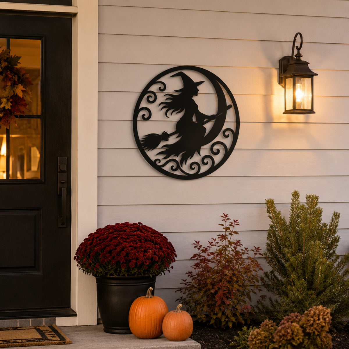 Black metal wall decor featuring a witch riding a broomstick around a crescent moon, mounted on a white siding exterior wall next to a lit lantern, with pumpkins and mums below.