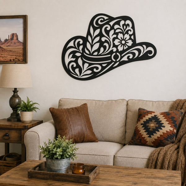 A black metal cowboy hat wall art piece with intricate white cutout floral patterns, mounted on a beige wall above a sofa in a styled living room.
