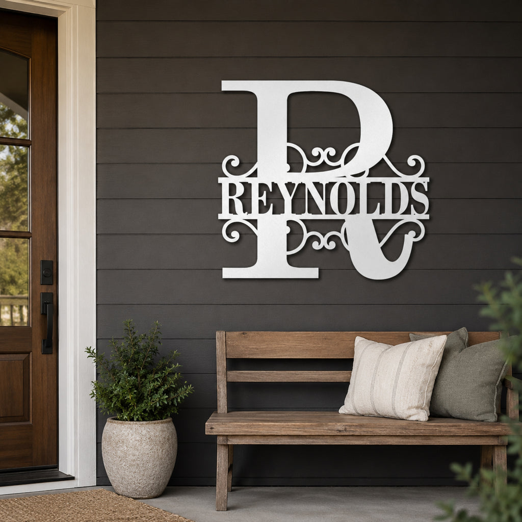 A large white decorative metal wall sign featuring a stylized letter R and the name REYNOLDS, mounted on a dark gray shiplap wall above a wooden bench with pillows in a front porch setting.