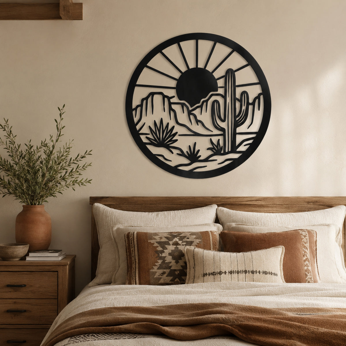 A circular black metal wall art piece featuring a desert landscape with a saguaro cactus, mountains, and a sun, displayed above a bed in a warm, neutral-toned bedroom.