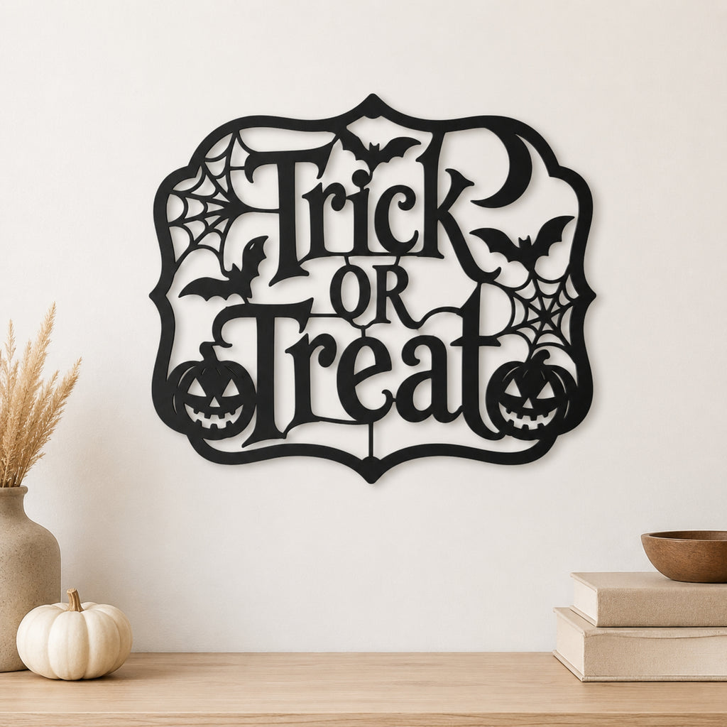 Black metal wall art with the words 'Trick or Treat' in a decorative cutout font, featuring Halloween motifs like bats, a crescent moon, spiderwebs, and jack-o'-lanterns, displayed on a light wall above a wooden surface with seasonal decor.