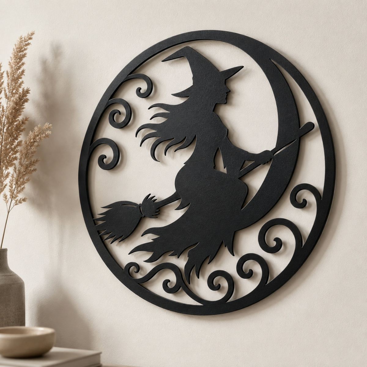 Black metal circular wall art featuring a witch silhouette riding a crescent moon on a broomstick, displayed on a beige wall with dried grass and a small vase in the background.