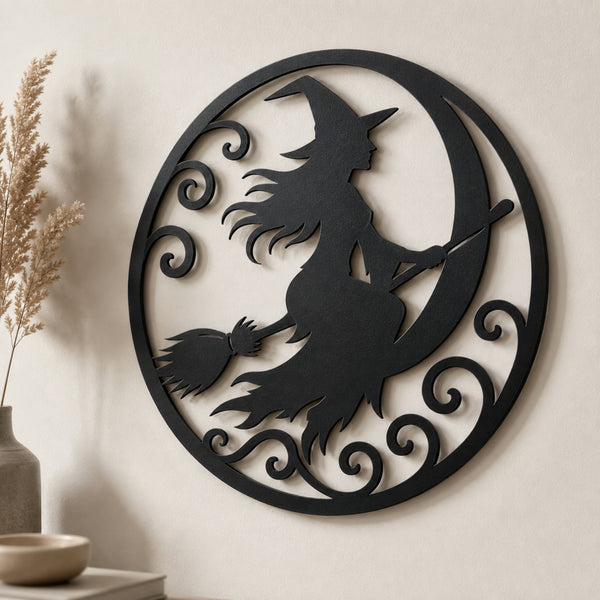 Black metal circular wall art featuring a witch silhouette riding a crescent moon on a broomstick, displayed on a beige wall with dried grass and a small vase in the background.