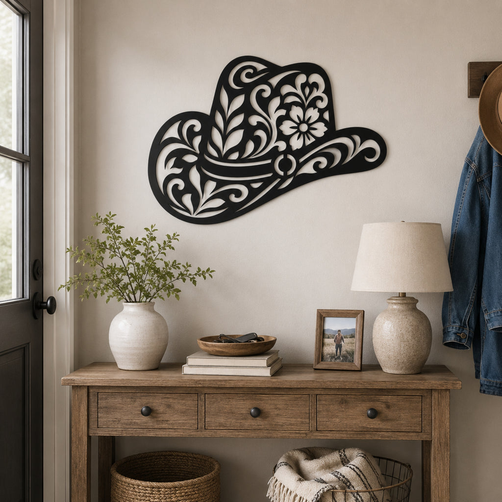A decorative black metal cowboy hat wall art piece with intricate white scrollwork and floral patterns, mounted on a beige wall above a rustic wooden console table styled with a vase of greenery, books, a lamp, and a framed photo.