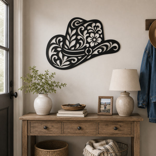 A decorative black metal cowboy hat wall art piece with intricate white scrollwork and floral patterns, mounted on a beige wall above a rustic wooden console table styled with a vase of greenery, books, a lamp, and a framed photo.