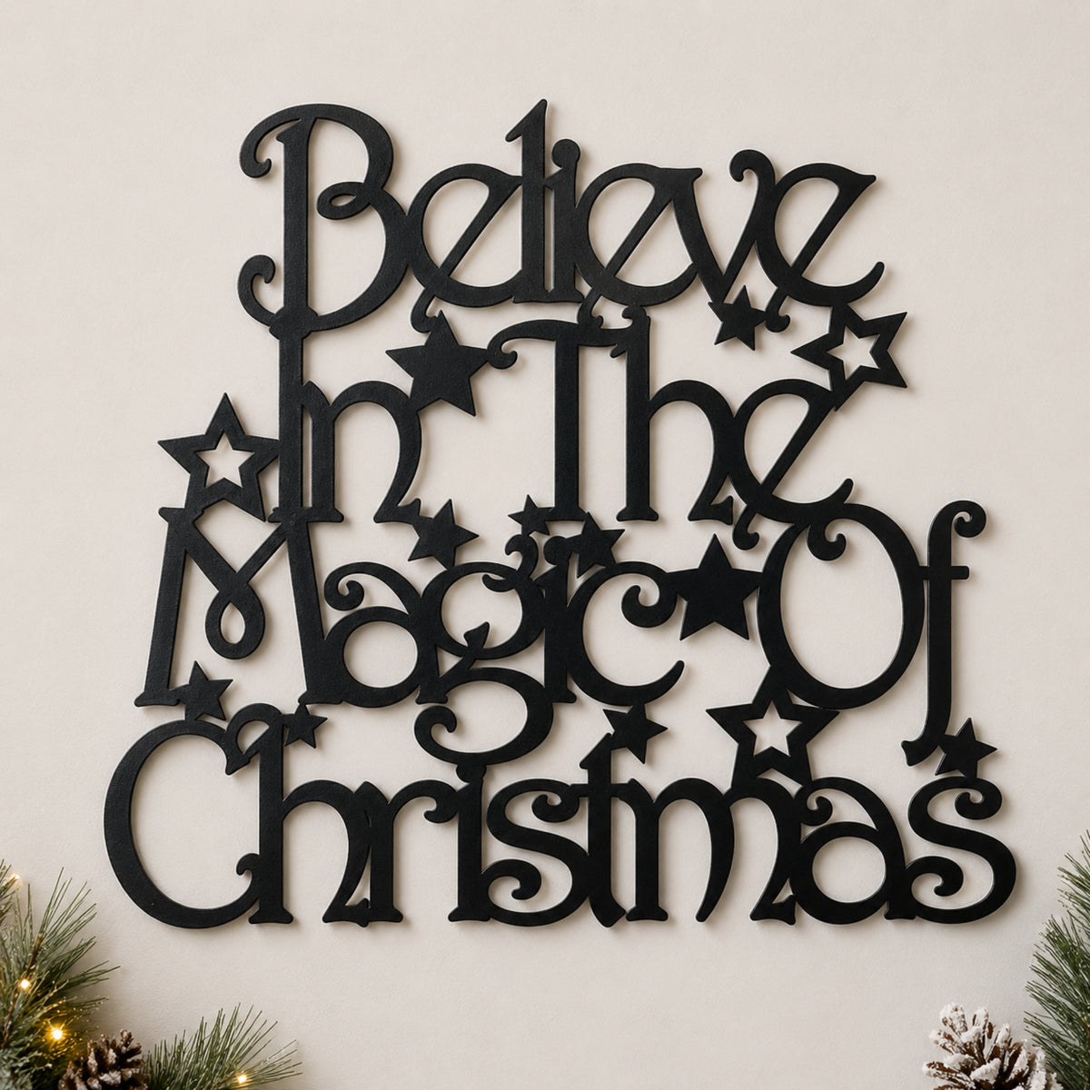 Black metal wall art with the phrase 'Believe in the Magic of Christmas' in decorative script and star shapes, mounted on a light beige wall with small pine and pinecone decorations at the bottom corners.