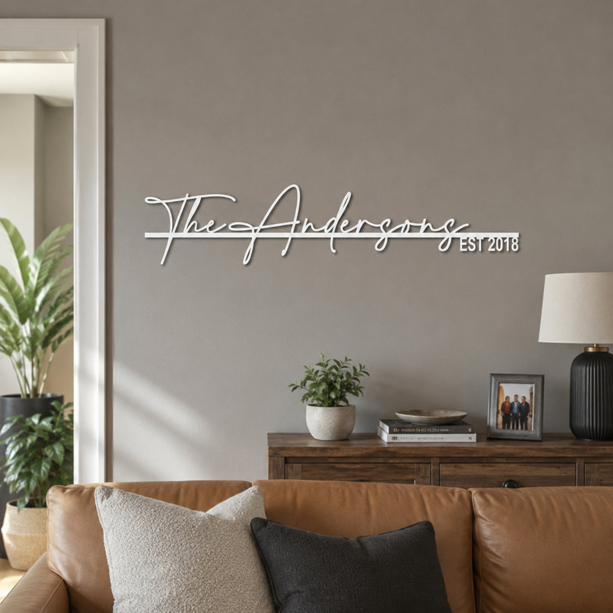 A white metal script wall sign reading 'The Andersons EST 2018' mounted on a gray living room wall above a wooden console table with a lamp, books, and a leather sofa.