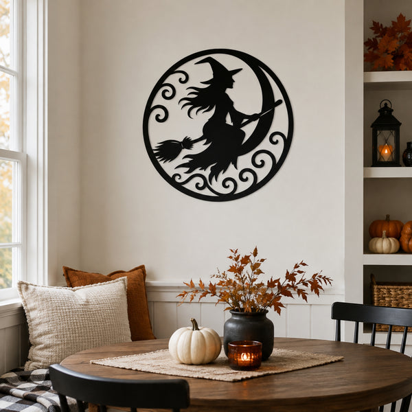 Black metal wall art featuring a witch riding a crescent moon with a broom, displayed on a beige wall in a styled dining room setting with autumn decor.