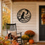 A black metal wall decor piece featuring a cat silhouette on a crescent moon and swirling branches, mounted on a beige house porch next to a black rocking chair and autumn flower arrangements.