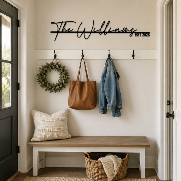 Custom black metal script wall sign reading 'The Williams EST 2025' mounted on a beige entryway wall above a white coat rack with a green wreath, tan tote bag, and blue denim jacket. A wooden bench with a cream pillow and a woven basket sit below.