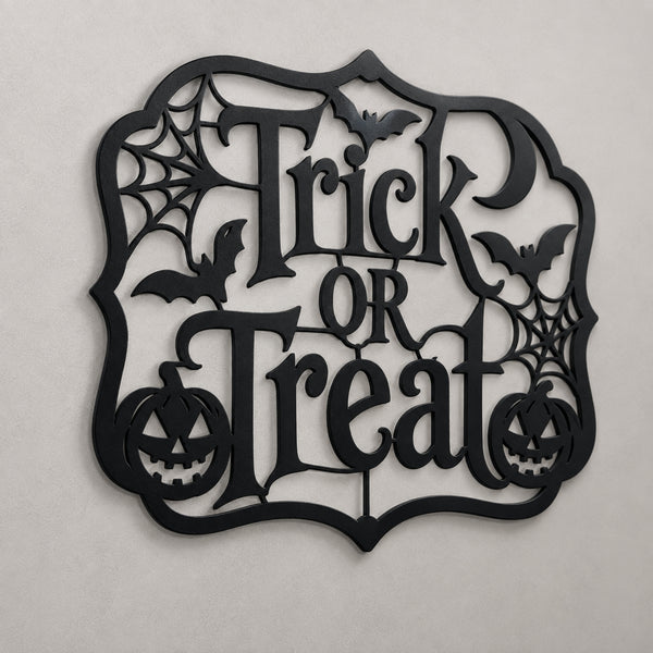 Black metal Halloween wall decor sign featuring the words 'Trick or Treat' in an ornate cutout font, surrounded by bats, spiderwebs, and jack-o'-lanterns, shown against a neutral light gray background.