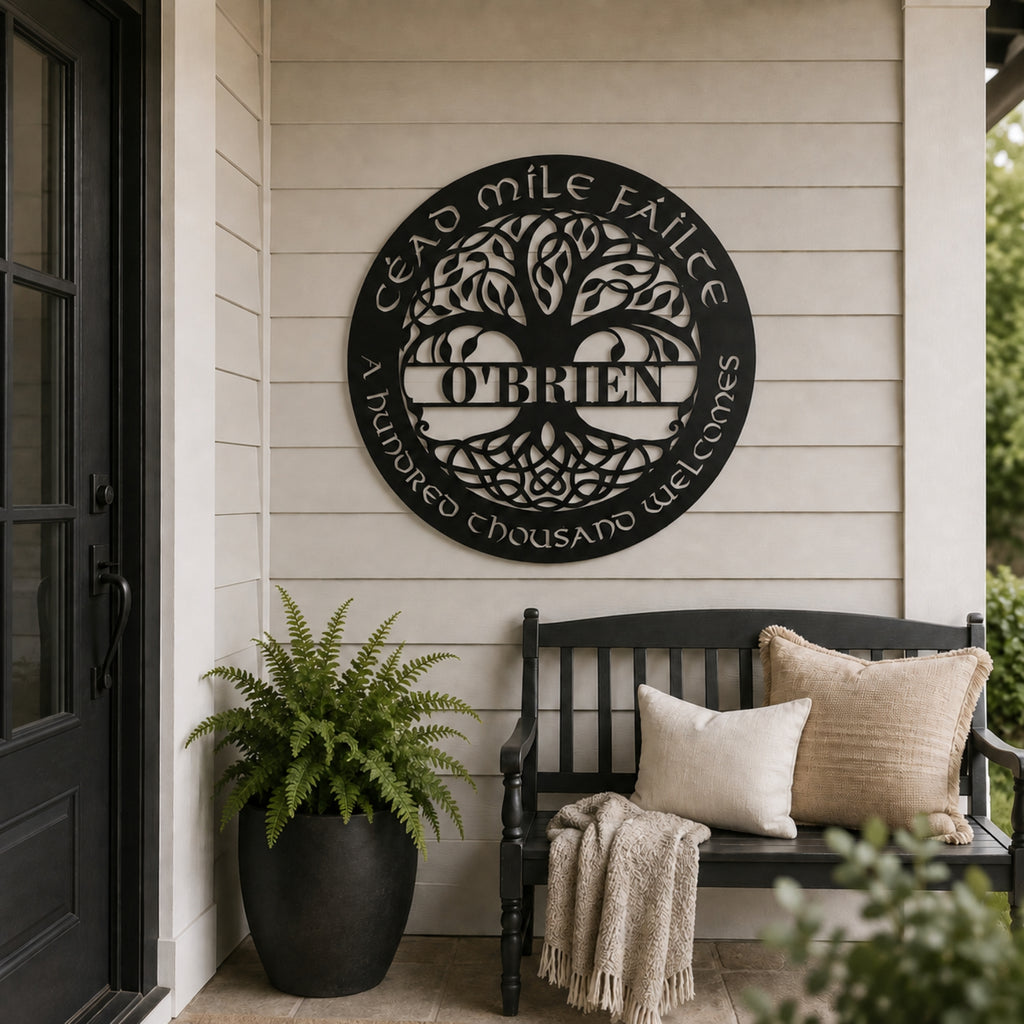 A circular black metal wall sign with a tree of life design and the name 'O'Brien' in the center, mounted on a beige house siding wall above a black bench with pillows and a throw blanket. A potted fern sits to the left.