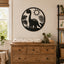 A black circular metal wall art piece featuring a dinosaur silhouette, sun, and palm leaves, displayed on a beige wall above a wooden dresser in a styled nursery setting.