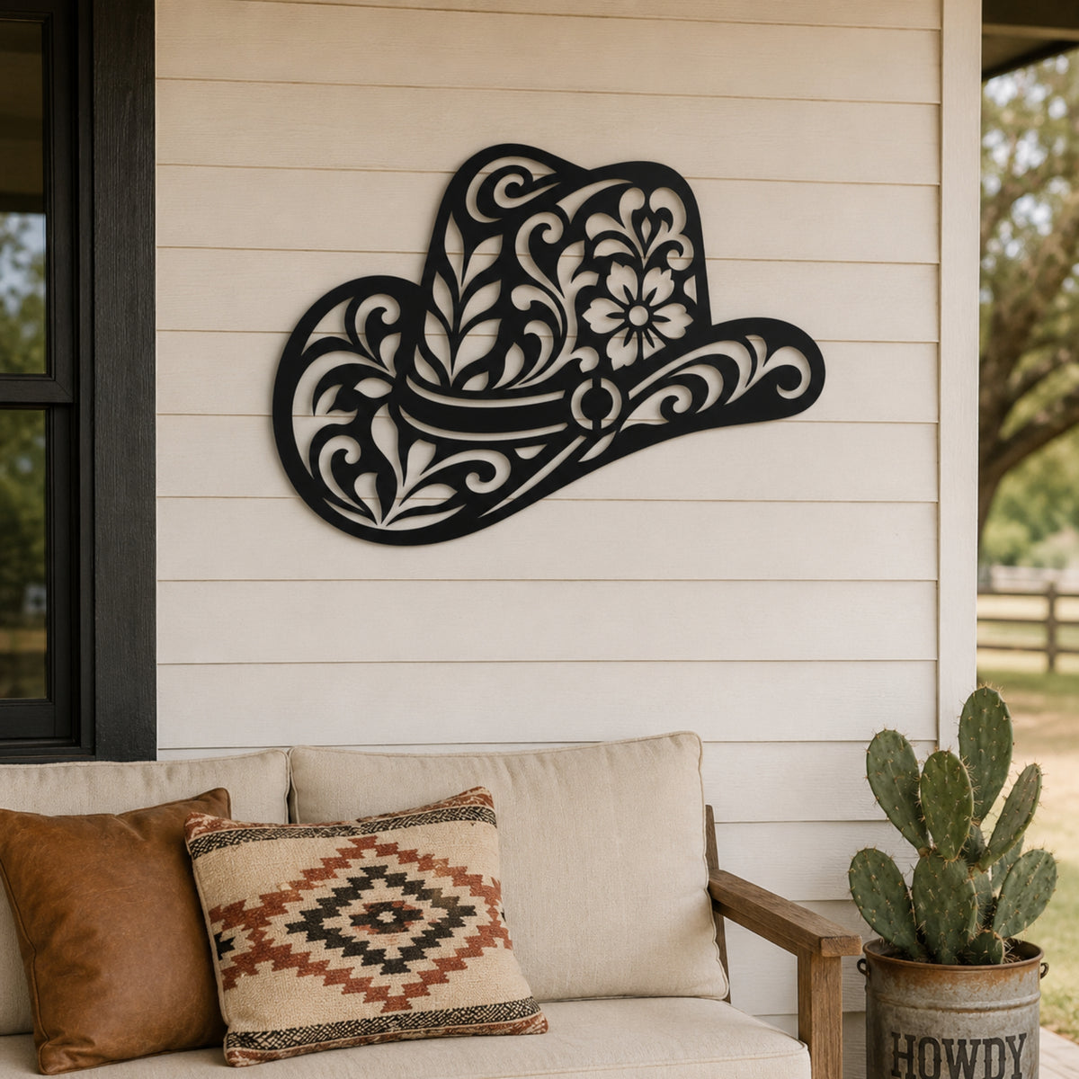 A decorative black metal wall sign shaped like a cowboy hat with intricate cutout floral and scroll patterns, mounted on a beige wood-paneled wall above a porch sofa with throw pillows and a potted cactus. Main image