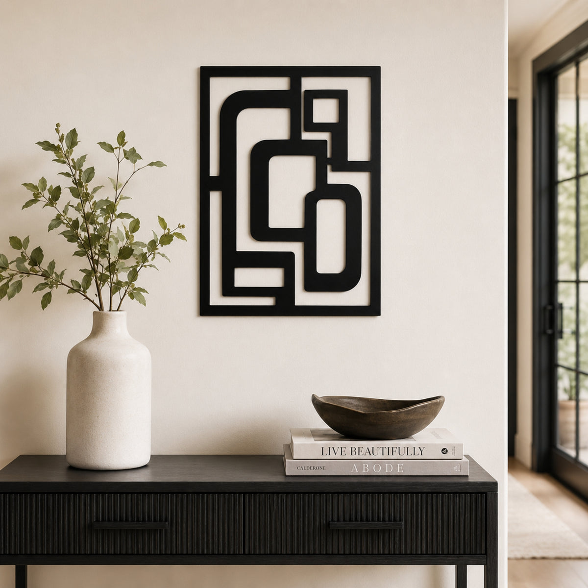 Black metal geometric wall art with an abstract frame design, displayed on a beige wall above a dark console table styled with a white vase and books. Main image