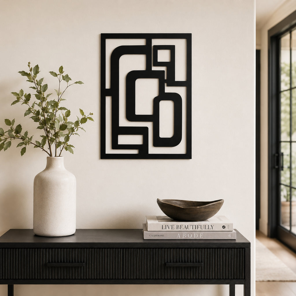 Black metal geometric wall art with an abstract frame design, displayed on a beige wall above a dark console table styled with a white vase and books.