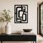 Black metal geometric wall art with an abstract frame design, displayed on a beige wall above a dark console table styled with a white vase and books.