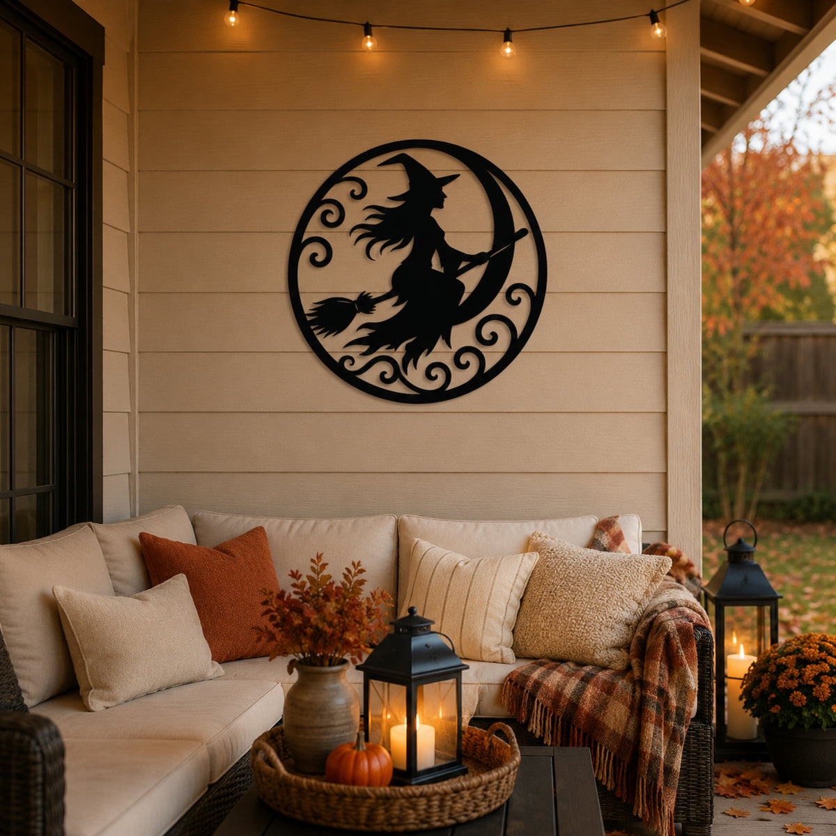 A black metal wall art piece featuring a witch riding a crescent moon with a broom, displayed on a beige porch wall above a cozy outdoor sofa decorated with pillows, a throw blanket, and fall decor. Main image