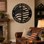 A round black metal wall sign featuring a central cross over an American flag design with the words 'LOVE OF COUNTRY' and 'FAITH IN GOD,' displayed on a beige wall in a living room above a brown leather sofa.