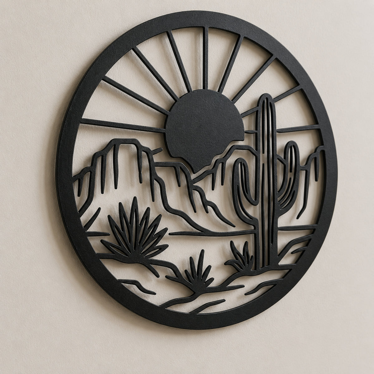 A circular black metal wall art piece featuring a desert landscape silhouette with a sun, mountains, saguaro cactus, and desert plants, mounted on a light beige wall.