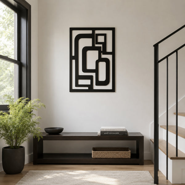 A black metal abstract geometric wall art piece hangs on a white wall above a dark wood console table in a modern entryway. A potted green plant sits on the left, and a black metal staircase railing is visible on the right.