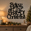 Black metal wall art with the phrase 'Believe in the Magic of Christmas' in an ornate script, mounted on a beige wall above a wooden console table decorated with a lamp, candle, pine garland, and a lit Christmas tree.