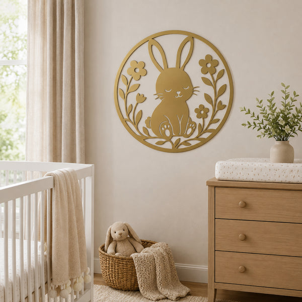 A circular gold metal wall art piece featuring a bunny and floral design, displayed on a beige wall in a styled nursery setting with a crib, dresser, and basket.