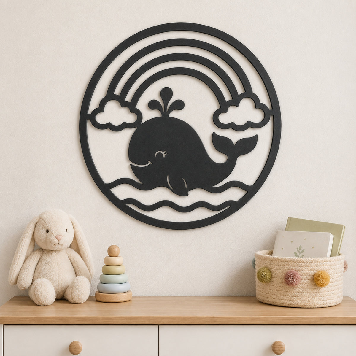 A black metal whale wall art piece with a rainbow and clouds, mounted on a beige wall above a wooden dresser styled with a plush bunny, a wooden stacking toy, and a woven basket.