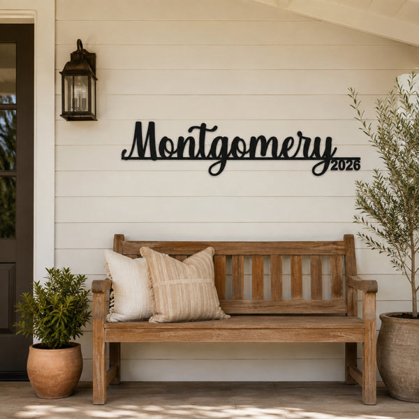 Black metal script wall sign reading 'Montgomery 2026' mounted on a beige house exterior wall above a wooden bench with decorative pillows, flanked by potted plants.
