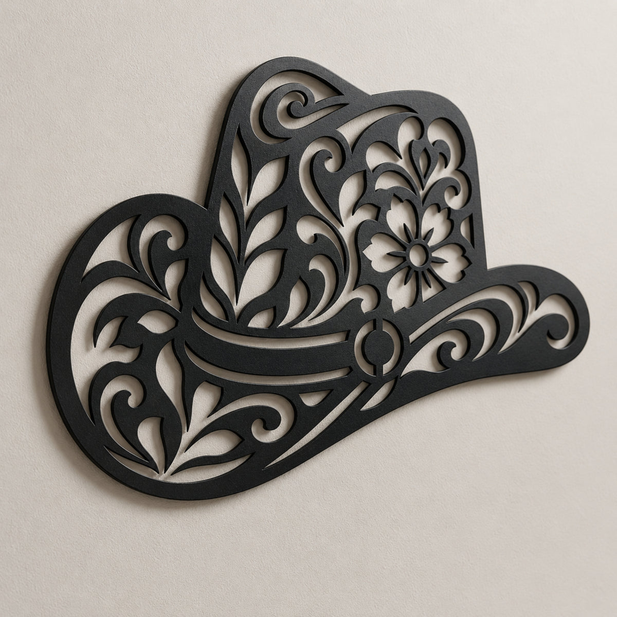 Black metal wall art shaped like a cowboy hat with ornate floral and scroll cutout details, mounted on a light beige textured wall.