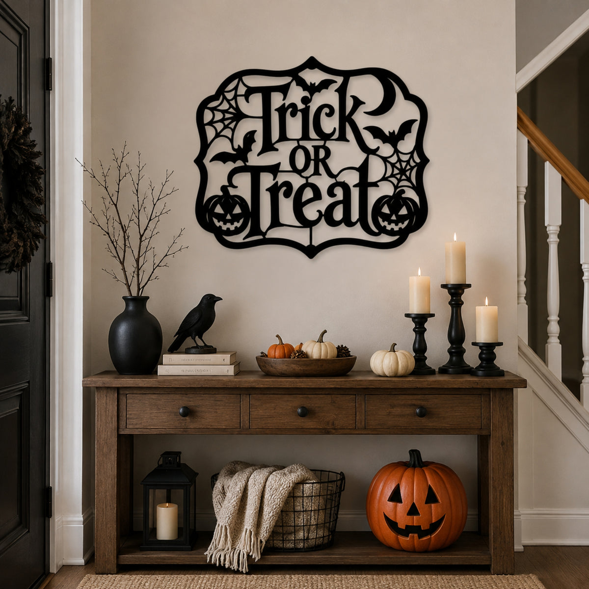 A black metal 'Trick or Treat' wall sign with bats, spiders, and jack-o'-lanterns is displayed on a beige wall above a wooden console table decorated for Halloween with candles, pumpkins, and a black bird figurine.