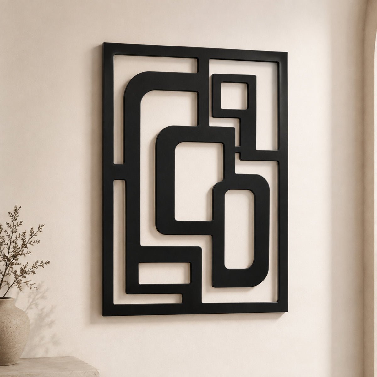 Black metal geometric wall art with an abstract maze-like pattern, mounted on a warm beige wall next to a small vase with dried branches.