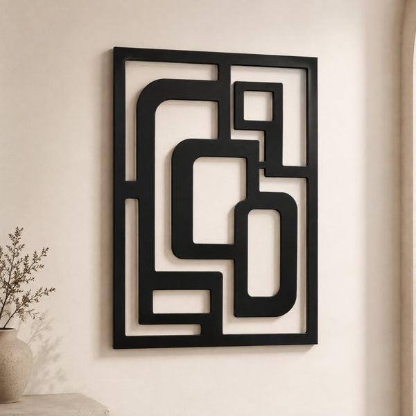 Black metal geometric wall art with an abstract maze-like pattern, mounted on a warm beige wall next to a small vase with dried branches.