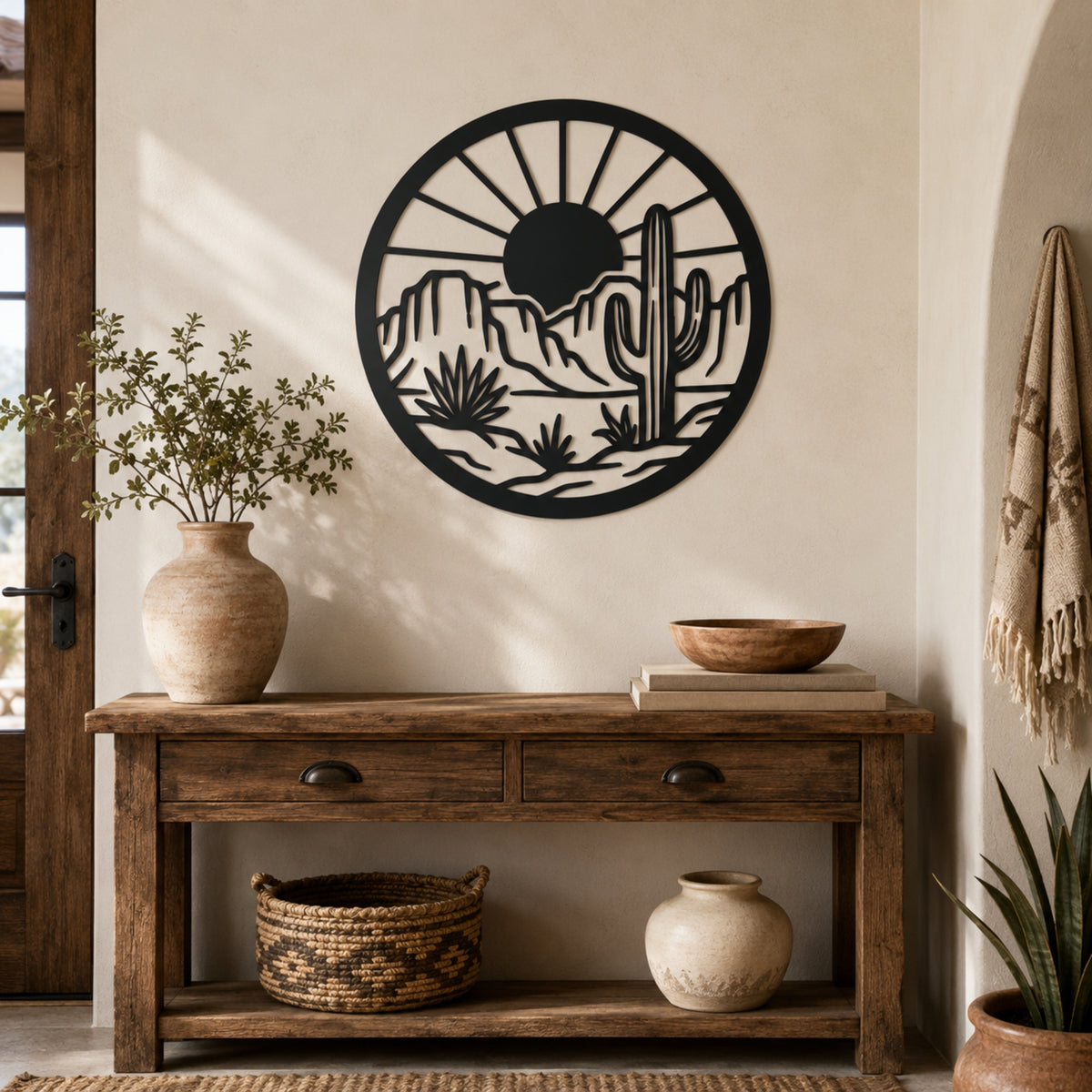 A circular black metal wall art piece featuring a desert landscape with a cactus and sun, mounted on a beige wall above a rustic wooden console table with decorative vases and baskets.