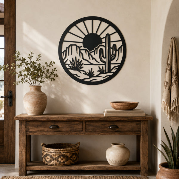 A circular black metal wall art piece featuring a desert landscape with a cactus and sun, mounted on a beige wall above a rustic wooden console table with decorative vases and baskets.