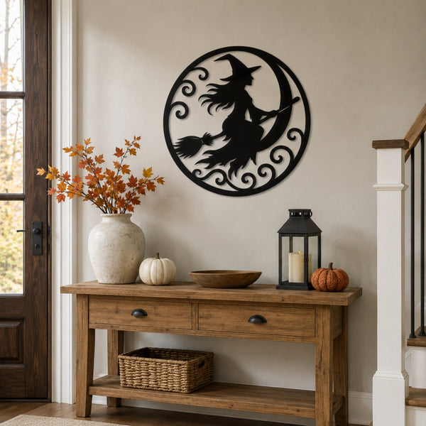 A black metal wall art piece featuring a witch riding a broomstick around a crescent moon, displayed on a beige wall above a rustic wooden console table decorated for autumn.