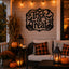 Black metal 'Trick or Treat' wall sign mounted on a beige wood-paneled porch wall, surrounded by fall decorations including string lights, pumpkins, lanterns, cornstalks, and a cushioned outdoor sofa with plaid pillows.